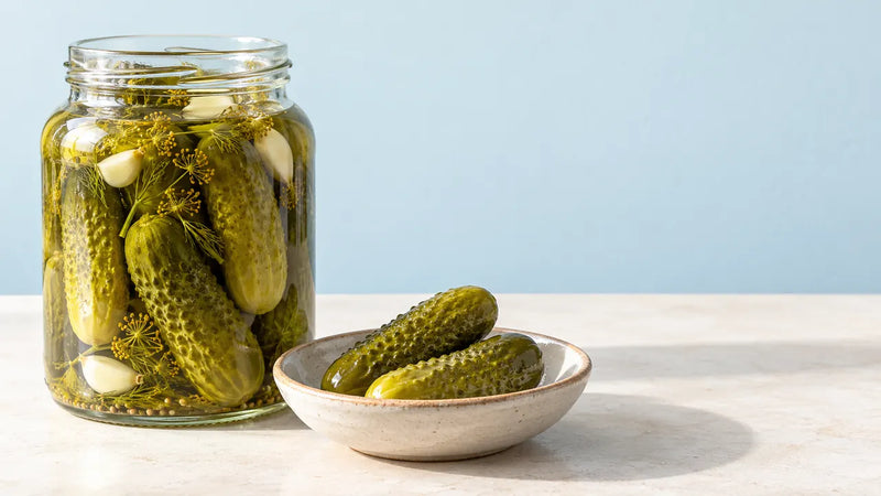 A glass jar of pickled cucumbers with dill and garlic beside a bowl of pickles.